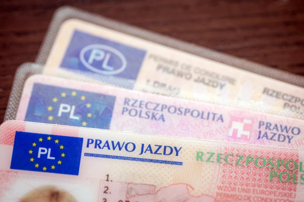 Polskie prawa jazdy różnych generacji ułożone w stos na drewnianym blacie, wyraźnie widoczne elementy graficzne oraz napisy identyfikujące dokumenty.