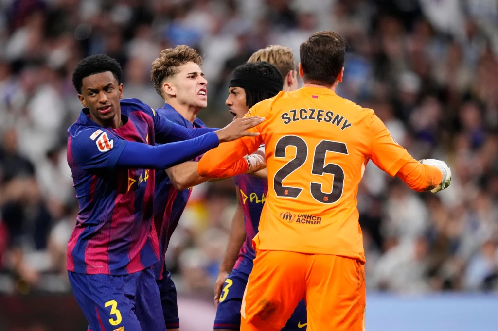 Przed momentem Wojciech Szczęsny obronił "jedenastkę". Barcelona wierzyła, że to przełomowy moment El Clasico Kilku piłkarzy w trakcie emocjonalnej wymiany podczas meczu piłkarskiego, zawodnik w pomarańczowym stroju z nazwiskiem Szczęsny stoi naprzeciw zawodników w pasiastych strojach, jeden z zawodników trzyma dłoń na ramieniu drugiego.