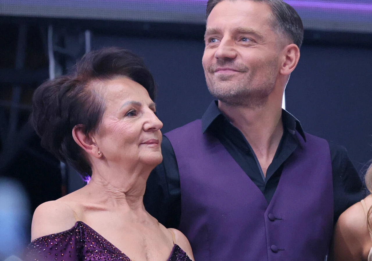 Ewa Rogacewicz, Marcin Rogacewicz Elegancka starsza kobieta i mężczyzna w fioletowych strojach stoją obok siebie w ciepłej atmosferze, oboje wyglądają na zadowolonych i skupionych, tło stanowi scena z fioletowym oświetleniem.