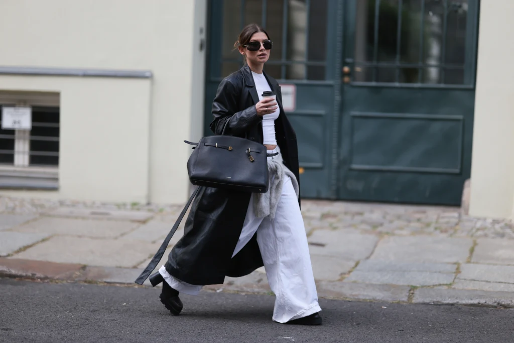 Stylizacja z białymi spodniami na jesień Kobieta w długim czarnym płaszczu, białych szerokich spodniach i okularach przeciwsłonecznych idzie ulicą z kawą w dłoni i dużą elegancką torebką.
