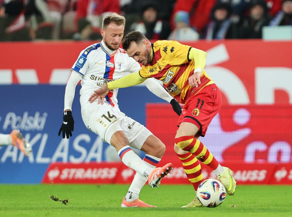 Jarosław Kubicki i Jesus Imaz podczas meczu Górnik Zabrze - Jagiellonia Białystok Dwóch piłkarzy walczy o piłkę podczas meczu, jeden zawodnik w białym stroju z niebiesko-czerwonymi detalami, drugi w żółto-czerwonym stroju, otoczeni żywo reagującą publicznością.