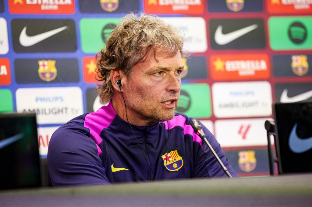 Marcus Sorg Trener piłkarski w stroju klubowym FC Barcelona siedzi na konferencji prasowej przed mikrofonem z słuchawką w uchu, w tle widoczne kolorowe tło z logo sponsorów i klubowymi barwami.