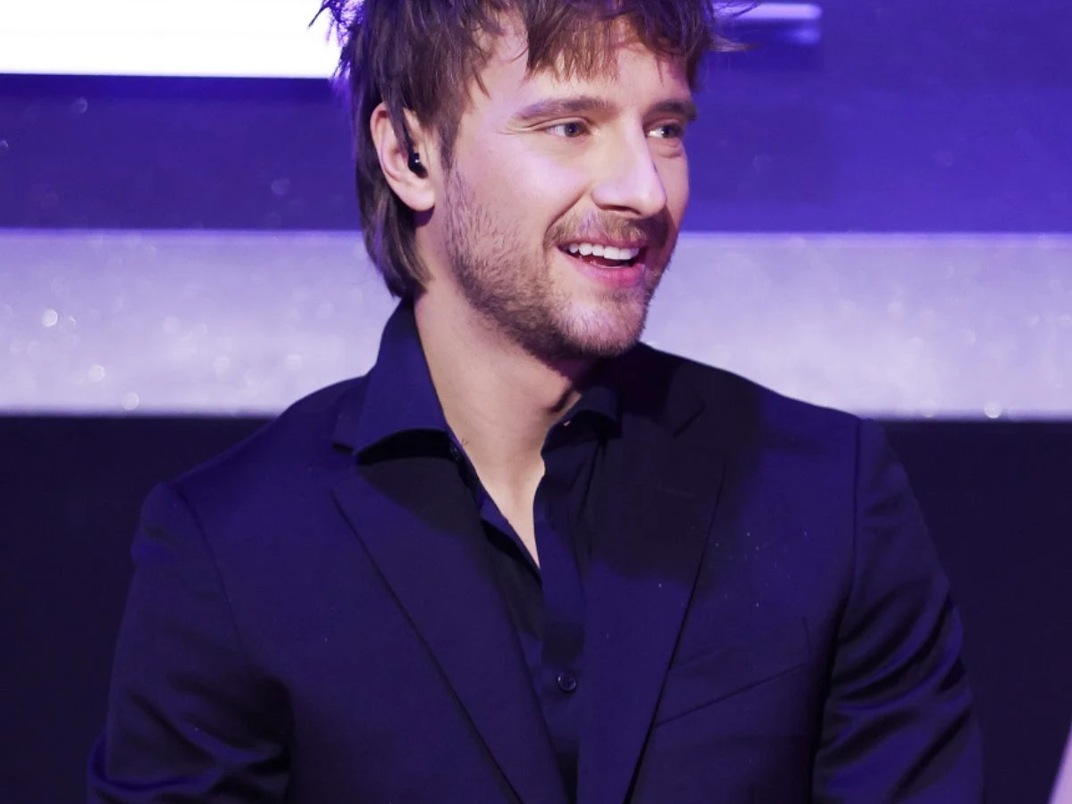 Maciej Musiał w "Tańcu z gwiazdami" Młody mężczyzna w garniturze z delikatnym zarostem i rozpuszczonymi włosami uśmiecha się, stojąc w oświetlonym, eleganckim otoczeniu.