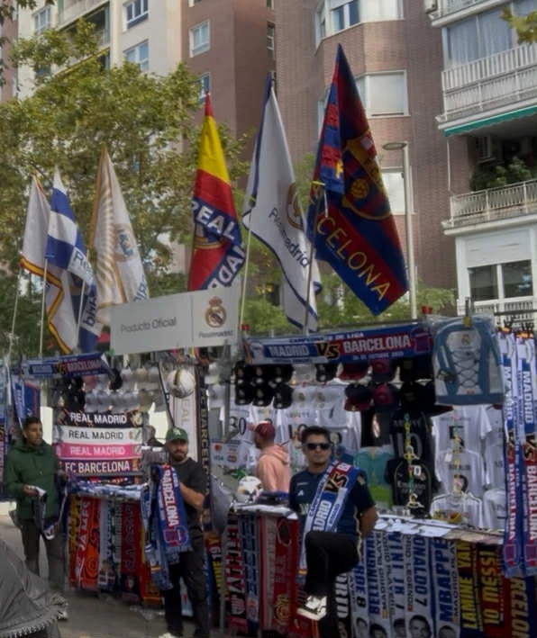 Stoisko z gadżetami piłkarskimi, głównie szalikami i flagami drużyn Real Madrid oraz Barcelona, ustawione na zewnątrz w przestrzeni miejskiej. W tle widoczne są bloki mieszkalne, kilku sprzedawców prezentuje oferowane produkty.