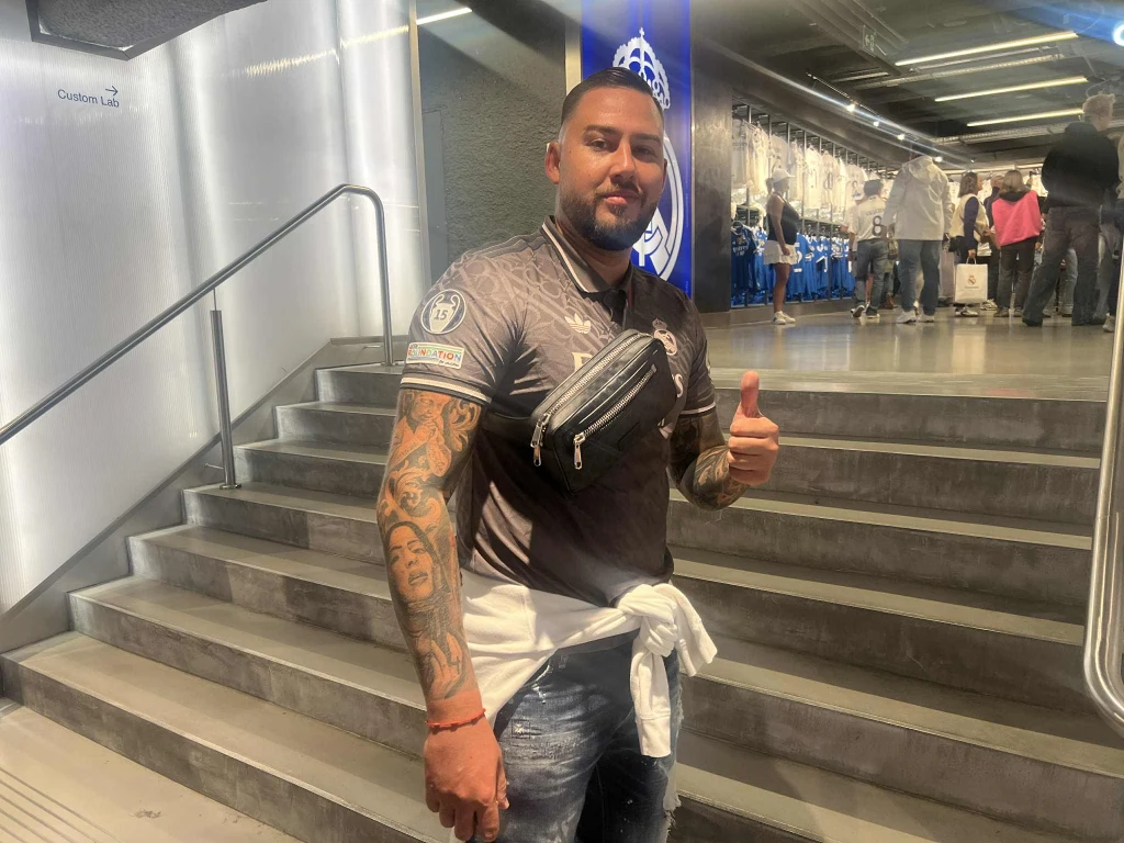 Uśmiechnięty mężczyzna z tatuażami na rękach pozuje na tle schodów, ubrany w sportową koszulkę i nerkę przewieszoną przez ramię, z kciukiem uniesionym w geście aprobaty, w tle osoby oraz nowoczesne wnętrze.