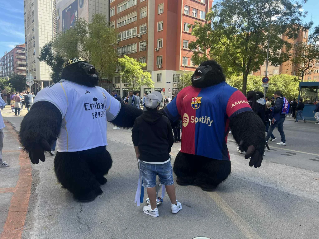 Dwie osoby w przebraniu wielkich goryli stoją naprzeciw siebie na środku ulicy, jeden w stroju piłkarskim Realu Madryt, drugi w koszulce FC Barcelony. Przed nimi stoi dziecko ubrana w sportowe ubrania. W tle widać przechodniów, drzewa i wysokie budynki.