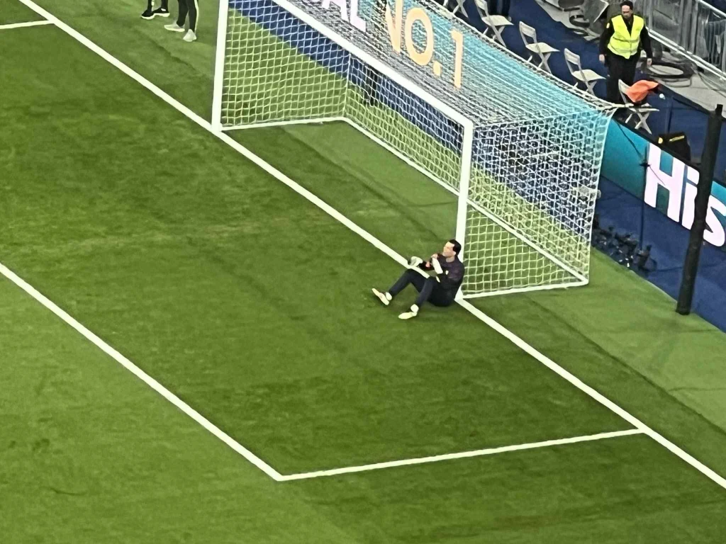 Bramkarz siedzący na murawie oparty o słupek bramki na stadionie piłkarskim, wokół widoczne puste krzesła i niewielu ludzi w tle.