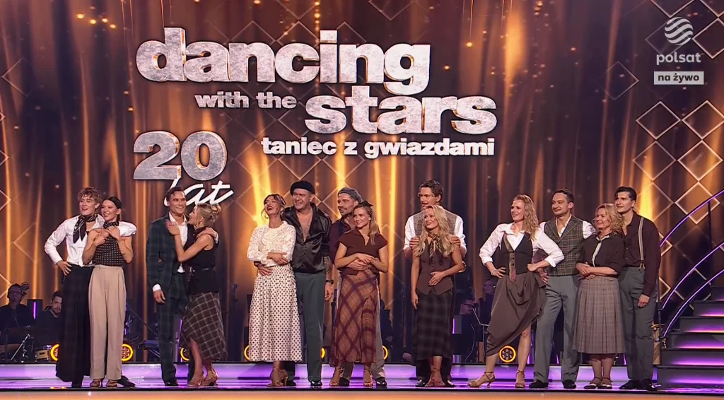 Potańcówka u Macieja Musiała - Taniec z Gwiazdami Uczestnicy programu tanecznego stojący grupowo na scenie przed dużym logo, ubrani w eleganckie stroje, na tle jasnego, widowiskowego oświetlenia.