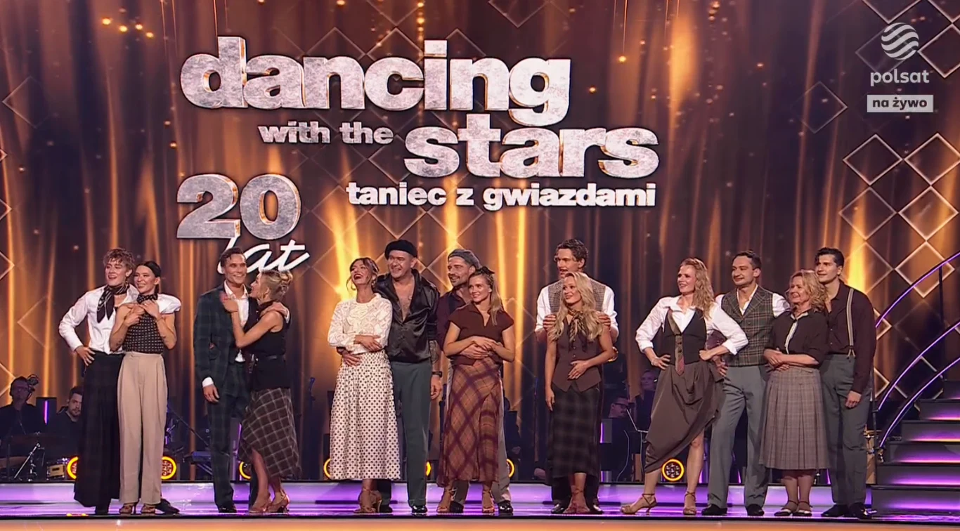 Uczestnicy programu tanecznego stojący grupowo na scenie przed dużym logo, ubrani w eleganckie stroje, na tle jasnego, widowiskowego oświetlenia.