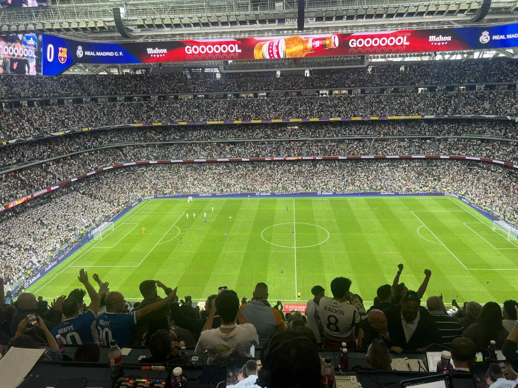 Widok na stadion piłkarski w czasie meczu, pełne trybuny kibiców, eksplozja radości po zdobyciu gola, na tablicach świetlnych napis „GOOOOOL” oraz herby drużyn Real Madryt i FC Barcelona.