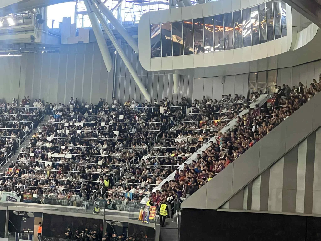 Tłum kibiców zajmuje trybuny nowoczesnego stadionu, większość osób skupiona na wydarzeniu sportowym, na dolnych poziomach widoczni stewardzi w żółtych kamizelkach dbający o bezpieczeństwo.