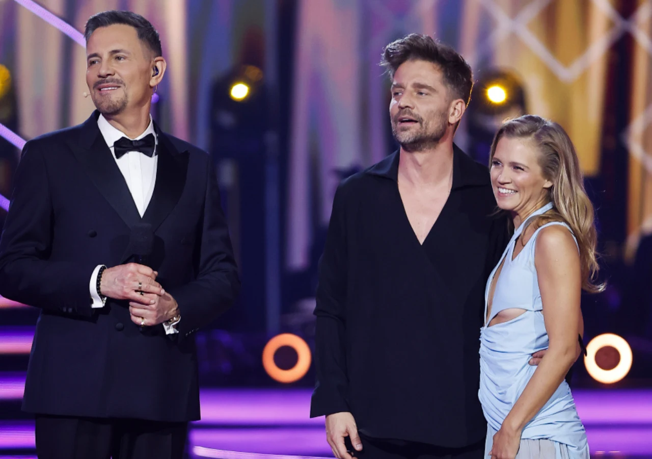 Trzy osoby na kolorowo oświetlonej scenie, mężczyzna w smokingu uśmiecha się i trzyma mikrofon, obok stoi para – mężczyzna w czarnej koszuli oraz kobieta w eleganckiej, jasnoniebieskiej sukni, obejmują się i również uśmiechają, atmosfera jest radosna i...