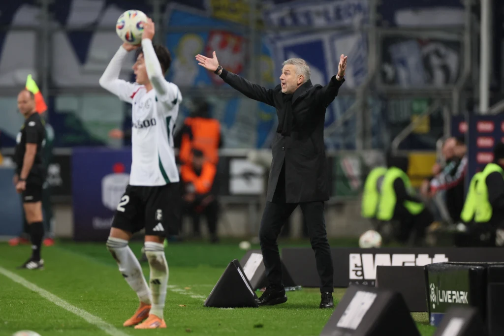 Poza Edwarda Iordanescu (w tle) okazała się doskonałą ilustracją hitu Ekstraklasy Trener w garniturze gestykuluje z szeroko rozłożonymi rękami przy linii bocznej boiska, podczas gdy zawodnik w białej koszulce przygotowuje się do wyrzutu z autu. W tle widoczny sędzia oraz fragment trybun.