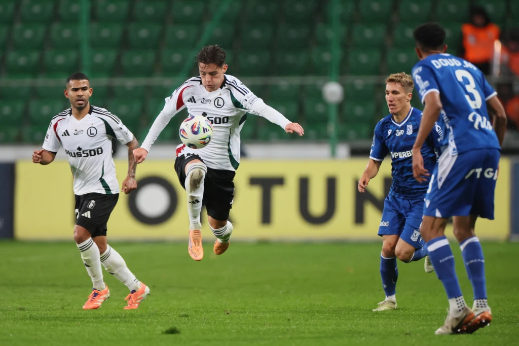 Kacper Urbański (przy piłce), tym razem najjaśniejsza postać w ekipie Legii Zawodnik w białej koszulce z logo klubu unosi się w powietrzu podczas odbicia piłki nożnej, otoczony przez innych piłkarzy ubranych w białe i niebieskie stroje. Akcja toczy się na zielonej murawie stadionu, w tle widoczna jest trybuna.