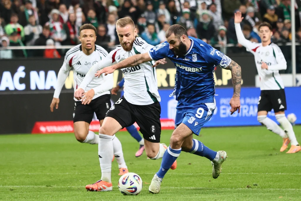 Mikael Ishak (nr 9) skutecznie zneutralizowany przez Rafała Augustyniaka Dwóch piłkarzy w trakcie dynamicznej walki o piłkę podczas meczu, otoczeni innymi zawodnikami i kibicami na trybunach.