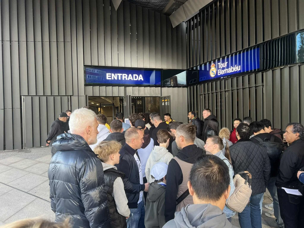 Kolejka do zwiedzania Bernabeu Tłum ludzi stojących w kolejce przed wejściem oznaczonym napisem 'ENTRADA' oraz szyldem 'Tour Bernabéu', większość osób jest ubrana w kurtki, wokół nowoczesna architektura budynku.