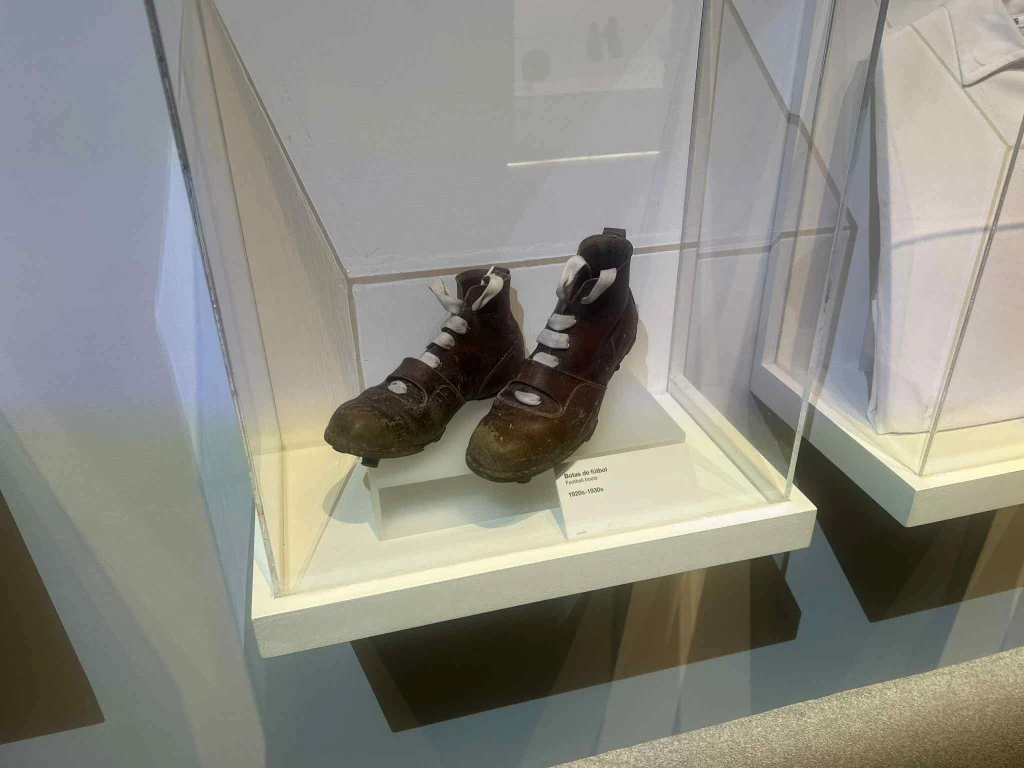 Buty piłkarskie pamiętające czasy sprzed 100 lat Stare sportowe buty z białymi sznurowadłami umieszczone w szklanej gablocie na ekspozycji muzealnej. Obuwie ma widoczne ślady zużycia i ciemne zabarwienie wynikające z intensywnego użytkowania.