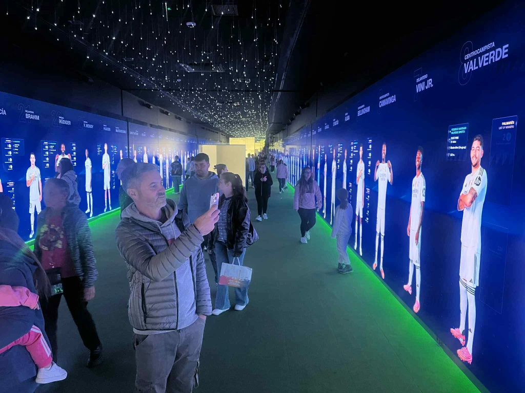 Zainscenizowana szatnia Realu Madryt Ludzie spacerują w korytarzu otoczonym ekranami z wizerunkami piłkarzy w strojach meczowych, na suficie diodowe światła przypominają gwiazdy, dominują kolory niebieski i zielony, część osób fotografuje ekrany.