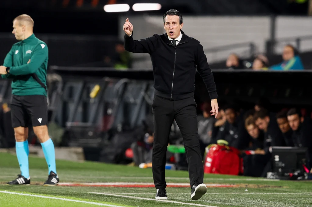 Unai Emery Trener piłkarski w eleganckim stroju gestykuluje na linii bocznej boiska, wskazując coś ręką. W tle widoczny sędzia oraz ławka rezerwowych z zawodnikami i sprzętem sportowym.