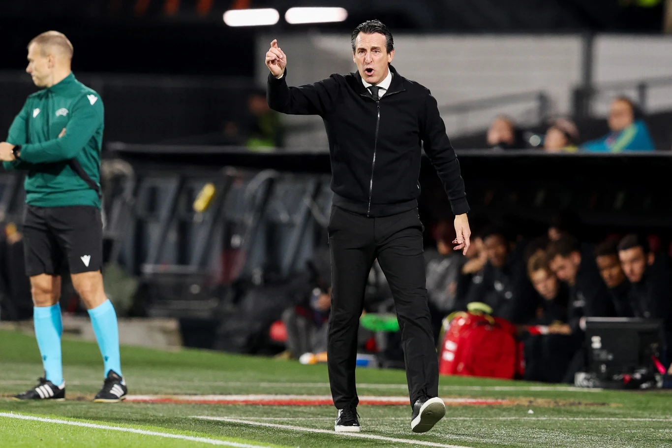 Trener piłkarski w eleganckim stroju gestykuluje na linii bocznej boiska, wskazując coś ręką. W tle widoczny sędzia oraz ławka rezerwowych z zawodnikami i sprzętem sportowym.