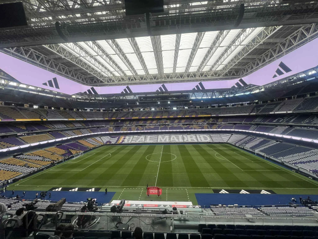 Widok na murawę Bernabeu Wnętrze dużego, nowoczesnego stadionu piłkarskiego z zielonym boiskiem i napisami Real Madrid na trybunach, dach stadionu częściowo przeszklony, siedziska w różnych kolorach, puste trybuny.