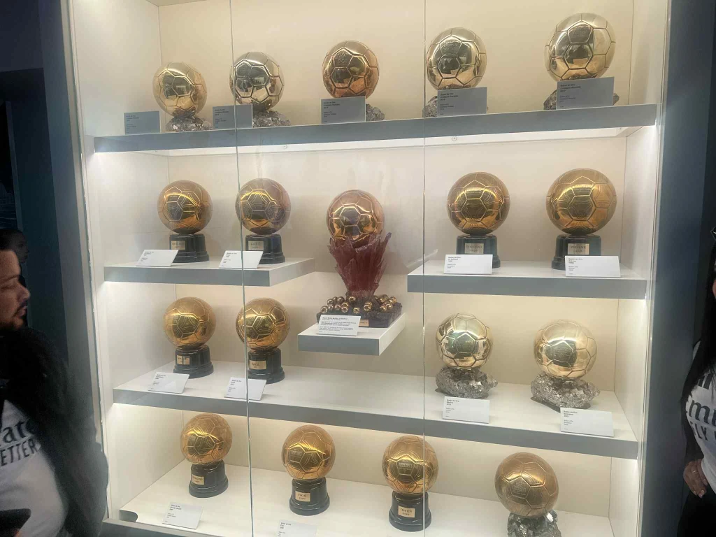 Gablota ze Złotymi Piłkami Gablota z wieloma Złotymi Piłkami oraz innymi trofeami piłkarskimi ustawionymi na kilku półkach, po bokach widoczne sylwetki zwiedzających.