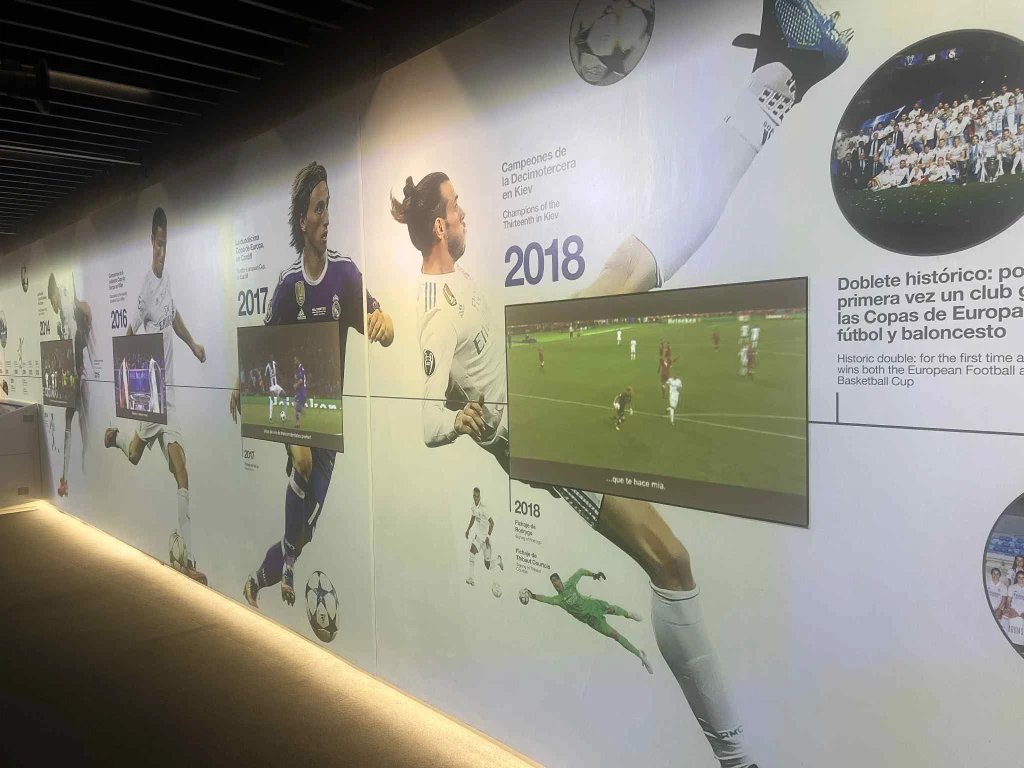 Multimedialna ściana z wygranymi finałami LM Ściana muzealna poświęcona sukcesom piłkarskim, z wkomponowanymi ekranami wyświetlającymi fragmenty meczów oraz grafikami i zdjęciami piłkarzy w akcji, rok po roku opisującymi dokonania drużyny.