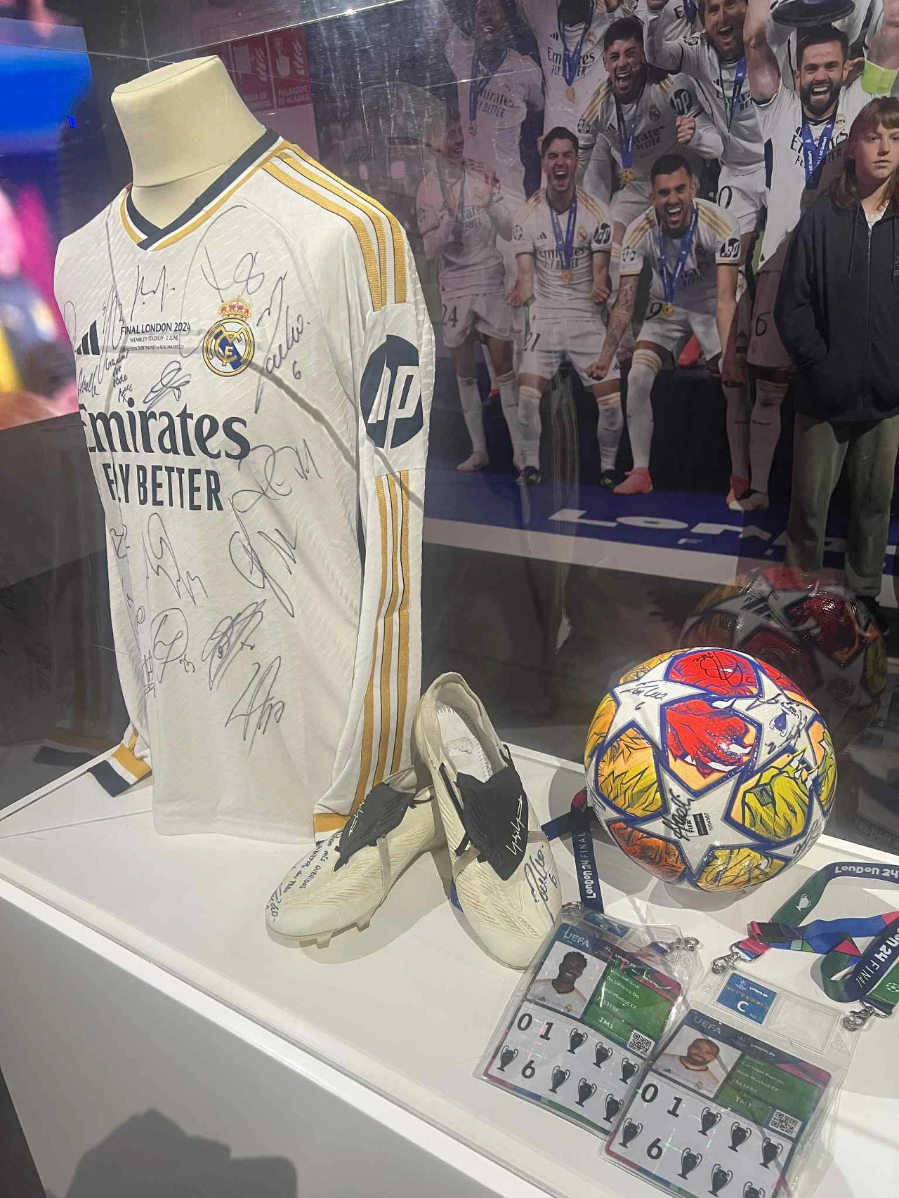 Koszulka z finału Ligi Mistrzów. Obok finałowe buty, piłka i akredytacje piłkarzy Koszulka piłkarska Realu Madryt z licznymi autografami zawodników, obok białe buty sportowe również podpisane, piłka z kolorowymi wzorami oraz dwie identyfikatory akredytacyjne. W tle drużyna świętująca sukces.
