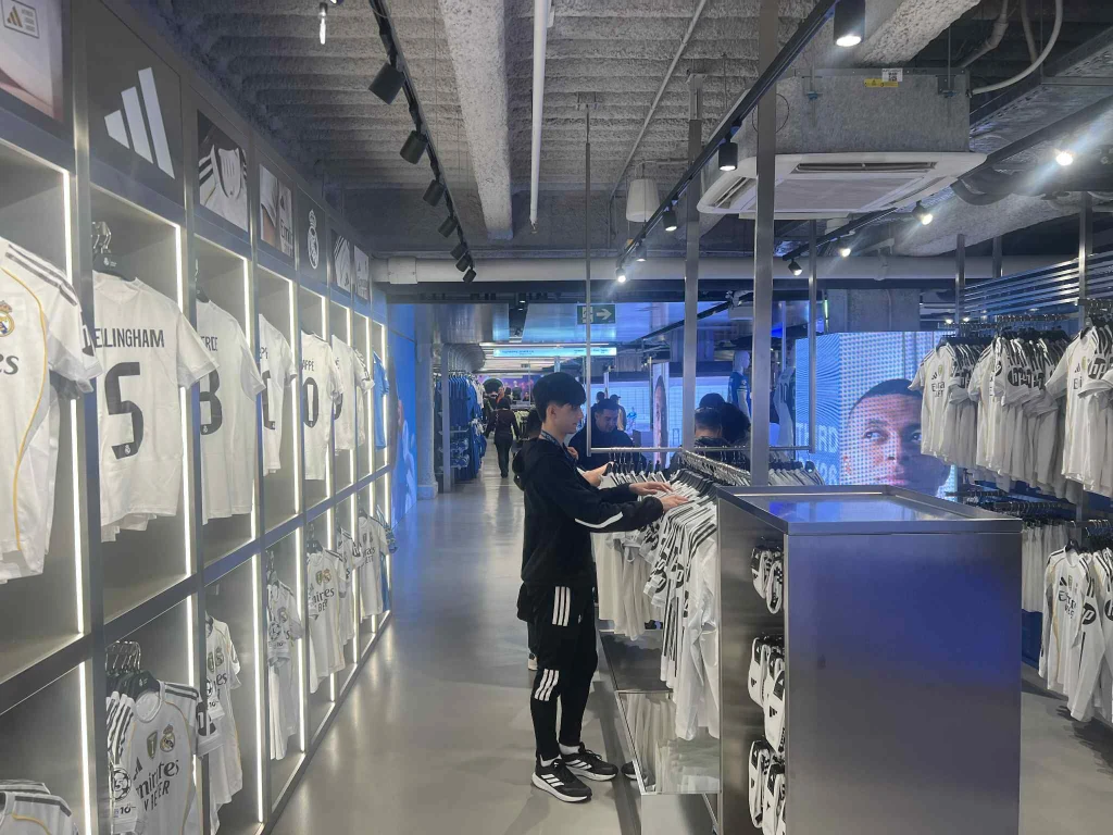 Klubowy sklep Osoba przeglądająca koszulki piłkarskie w oficjalnym sklepie, ściany wypełnione białymi trykotami Realu Madryt z nazwiskami zawodników, wnętrze nowoczesnego sklepu sportowego z jasnym oświetleniem.