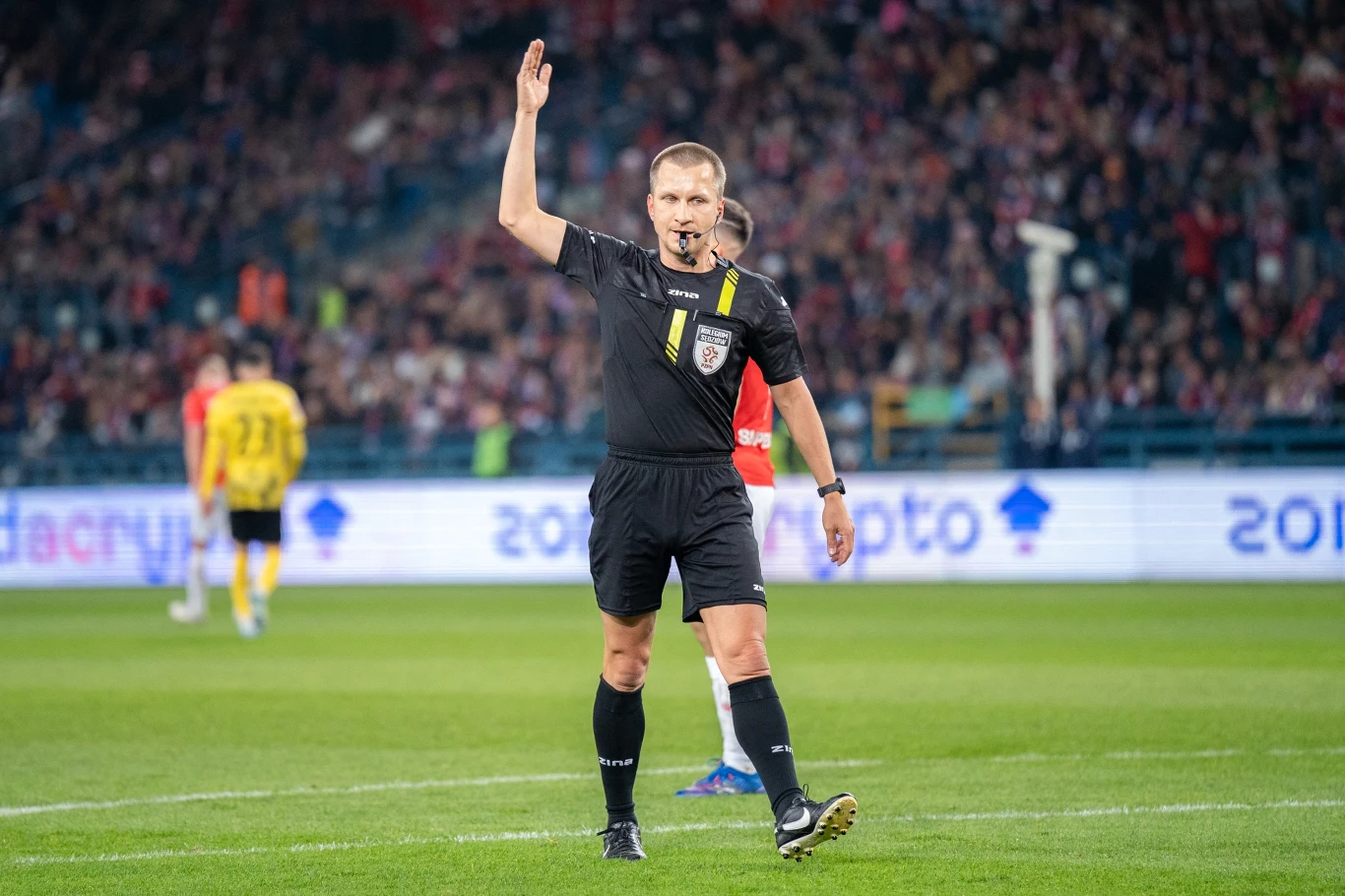 Sędzia piłkarski w czarnej koszulce z podniesioną ręką gwiżdże na boisku w trakcie meczu, w tle widoczni piłkarze i trybuny wypełnione kibicami.