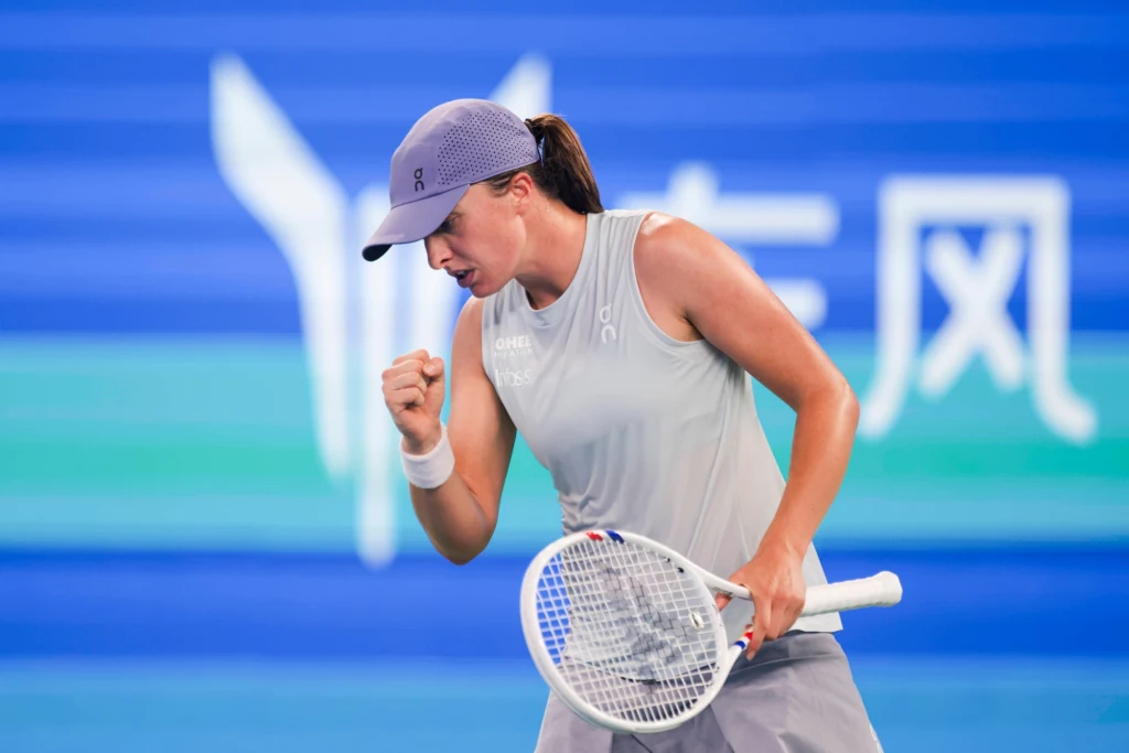 Tenisistka w sportowym stroju wykonuje gest triumfu, trzymając rakietę tenisową na tle niebieskiego kortu i wyraźnych białych napisów w tle.