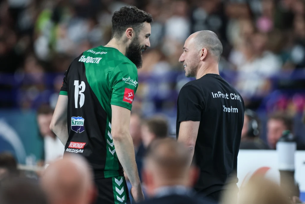 Krzysztof Andrzejewski (z prawej) i Łukasz Swodczyk Dwóch mężczyzn w sportowych strojach rozmawia ze sobą podczas wydarzenia sportowego, otoczeni przez rozmytą widownię. Jeden z nich ubrany jest w zielono-czarną koszulkę z numerem 13, drugi w czarną koszulkę z napisem na plecach.