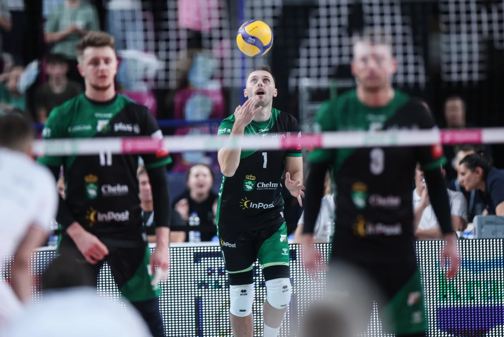 Siatkarze InPost ChKS Chełm Zawodnik drużyny siatkarskiej w zielono-czarnym stroju przygotowujący się do rozegrania piłki, wokół inni gracze i fragment siatki, w tle widoczni kibice i trybuny.
