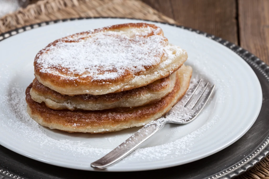 Trzy puszyste placki drożdżowe obsypane cukrem pudrem ułożone na białym talerzu, obok srebrny widelec, całość na dekoracyjnej podkładce.