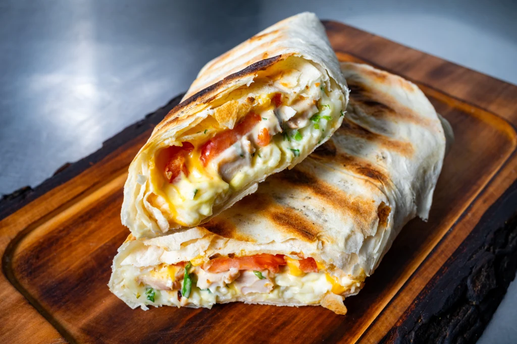 Dwa kawałki grillowanego wrapa z tortilli ułożone na desce drewnianej, wypełnione składnikami takimi jak kurczak, ser, jajko, sałata oraz pomidor, widoczne apetyczne nadzienie.