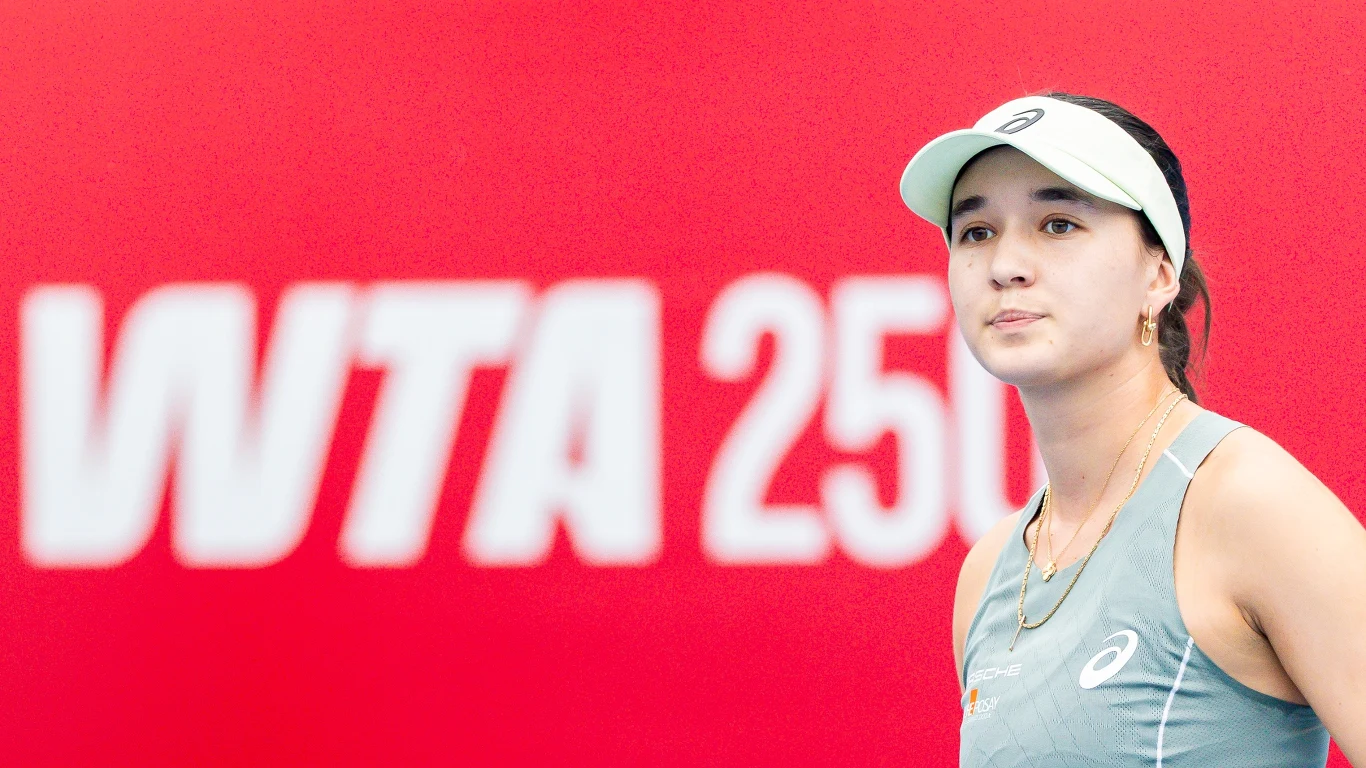 Tenisistka w sportowym stroju i białej czapce z daszkiem stoi na tle czerwonej ściany z dużym, białym napisem WTA 250.