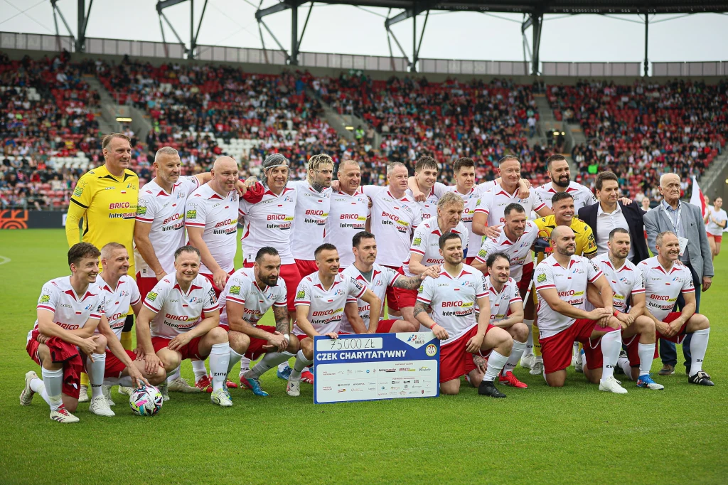 Grupa mężczyzn w piłkarskich strojach ustawia się do wspólnego zdjęcia na murawie stadionu. Wśród nich kilka osób w ubraniach cywilnych, wszyscy uśmiechnięci, trzymają duży symboliczny czek charytatywny. Na trybunach widoczni kibice.