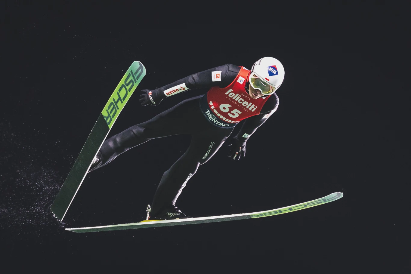 Kamil Stoch na skoczni zimowych igrzysk olimpijskich 2026 w Predazzo Skoczek narciarski w locie na tle ciemnego nieba, ubrany w profesjonalny kombinezon i kask, dynamiczna sylwetka podkreśla intensywność sportowej rywalizacji.