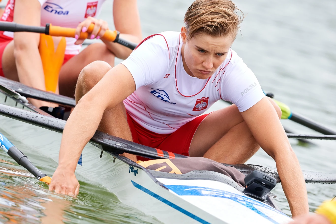 Katarzyna Zillmann Wioślarz w sportowej koszulce z orzełkiem na tle biało-czerwonym podczas wyścigu na wodzie, intensywnie skupiony nad ruchem wiosła, w tle pozostali członkowie osady.