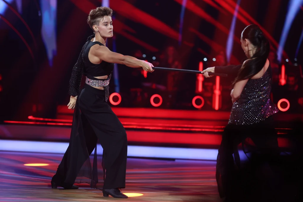 Dwie tancerki w trakcie dynamicznego występu z rekwizytem, eleganckie kostiumy i intensywne światła sceniczne w tle, ruch uchwycony w momencie przeciągania laski.