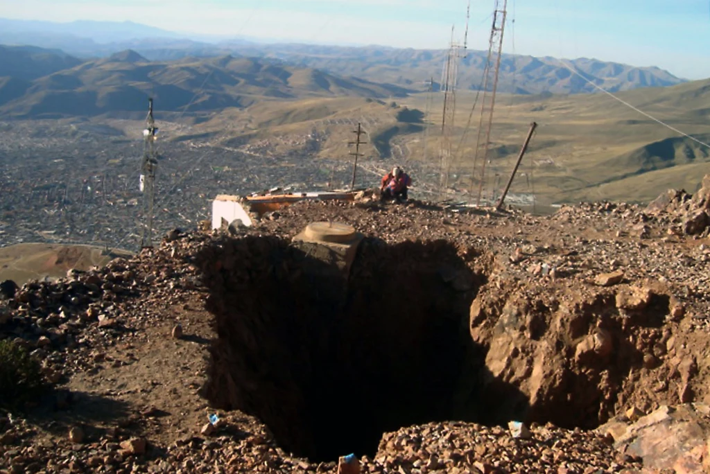 Wielka dziura w ziemi na skalistym wzgórzu, obok której ustawiono metalowe maszty i przewody. W oddali rozpościera się panorama miasta i górzystego krajobrazu. Na skraju dziury znajduje się osoba siedząca na kamieniach.