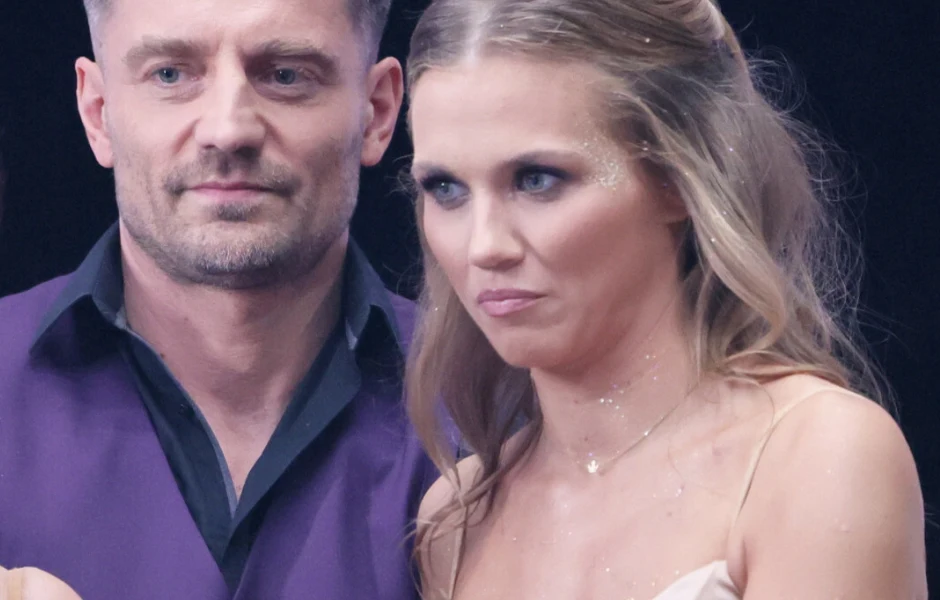 Para elegancko ubranych osób stoi blisko siebie, kobieta o długich, falowanych włosach w jasnej sukni z delikatnym makijażem i brokatem na ramieniu oraz mężczyzna w fioletowej kamizelce i ciemnej koszuli, oboje patrzą przed siebie.