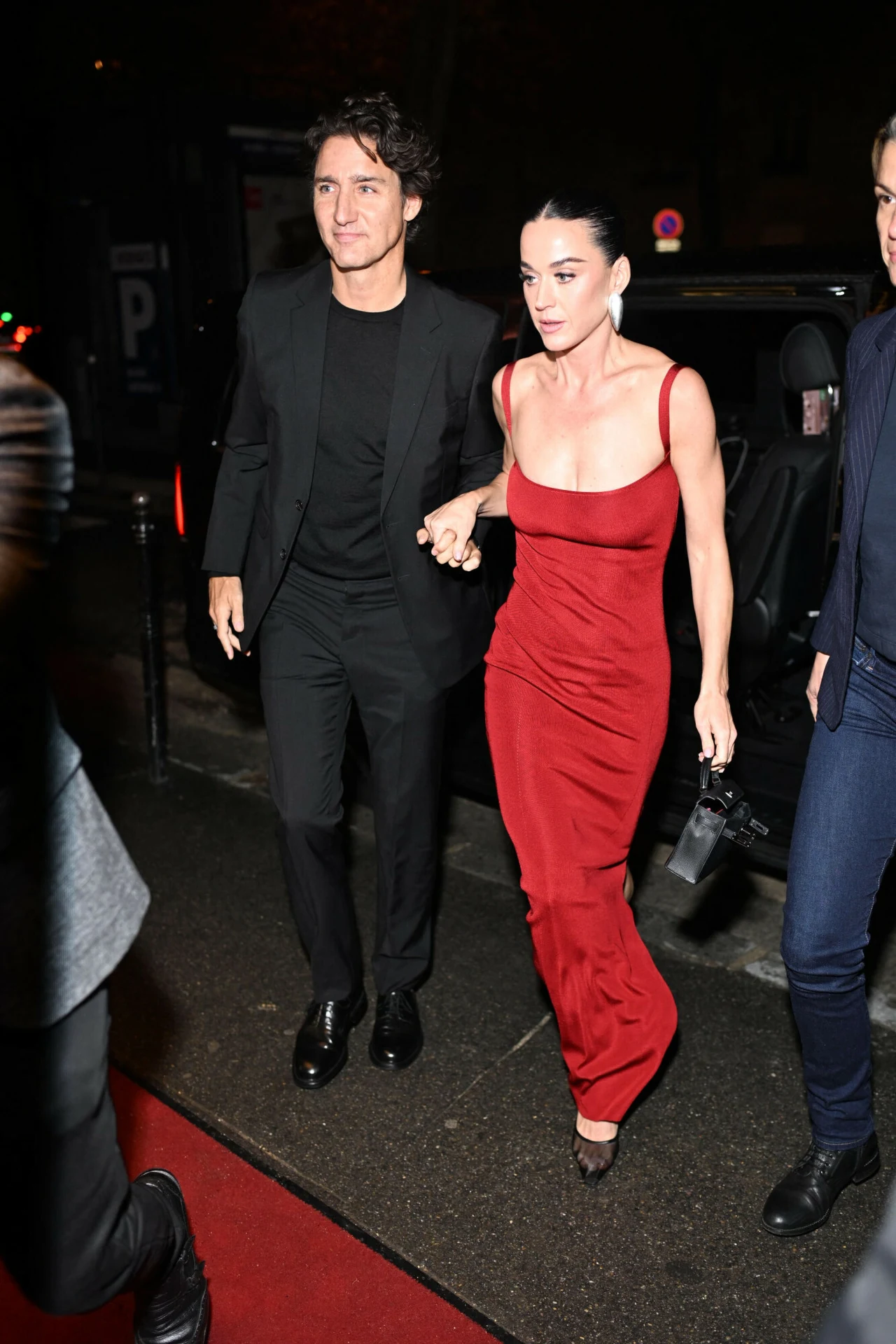 Katy Perry i Justin Trudeau są parą? Elegancka kobieta w długiej, czerwonej sukni i mężczyzna w czarnym garniturze idą razem po czerwonym dywanie nocą, trzymając się za ręce. Tłum i samochody w tle, atmosfera formalnego wydarzenia.