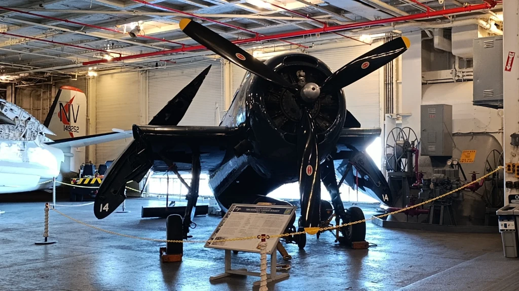 W czasie wojny na Pacyfiku na USS Hornet mógł bazować do około 90 samolotów. Wśród nich były myśliwce F6F Hellcat (później F4U Corsair), bombowce nurkujące SB2C Helldiver oraz torpedowce TBF-1 Avenger. Na zdjęciu jeden z zachowanych Hellcatów w hangarze lotniskowca Czarny, zabytkowy samolot wojskowy z śmigłem i charakterystycznym malowaniem prezentowany w hangarze muzealnym, otoczony tablicą informacyjną oraz barierkami ograniczającymi dostęp, w tle widoczny fragment drugiego samolotu.