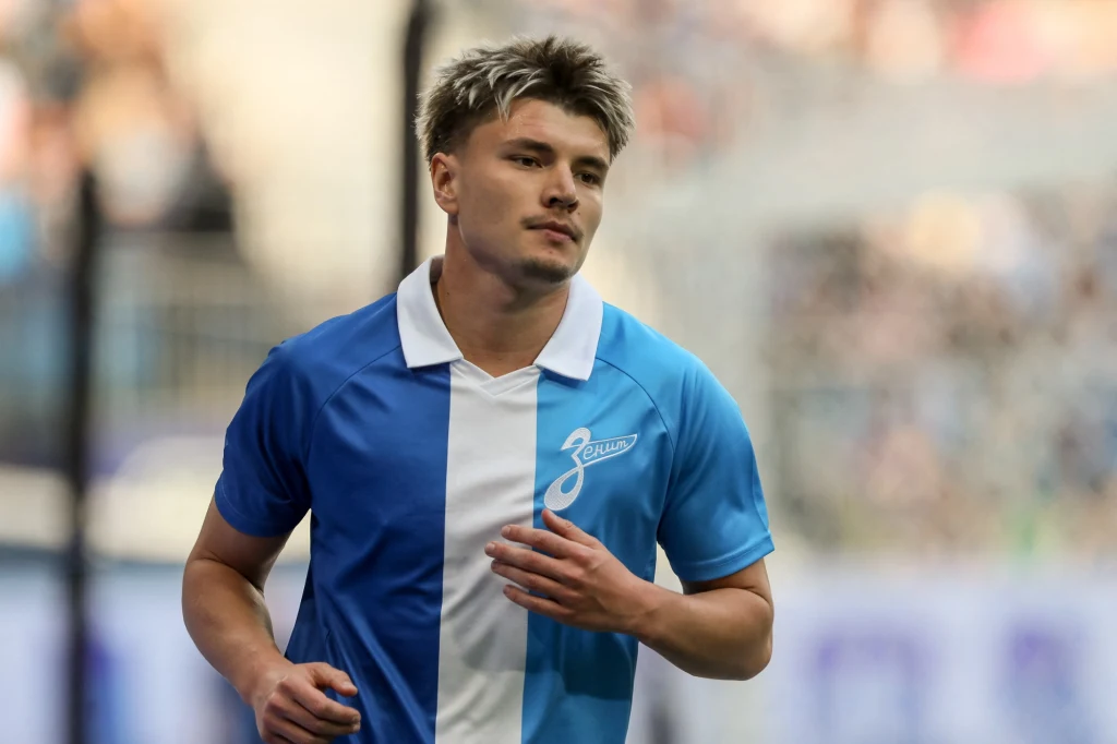 Andriej Mostowoj Młody mężczyzna w sportowej koszulce biało-niebieskiej z emblematem klubu Zenit na tle rozmytej publiczności, podczas aktywności sportowej na stadionie.