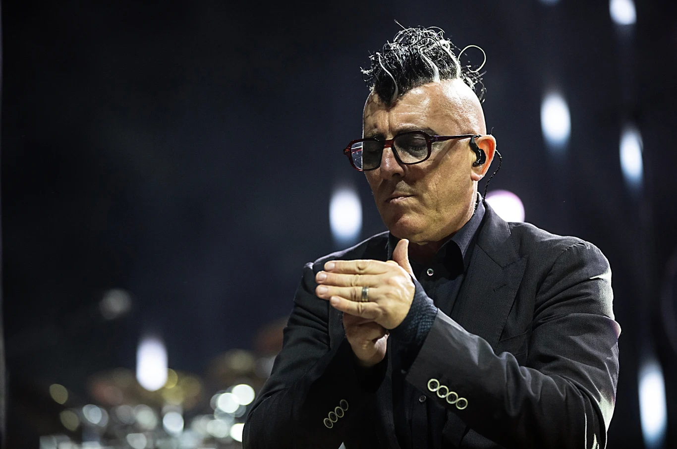 Maynard James Keenan powróci do Polski z grupą A Perfect Circle Mężczyzna w ciemnych okularach i czarnym garniturze z rozjaśnioną fryzurą typu irokez stoi na scenie i klaszcze w dłonie. W tle widoczne rozmyte światła.