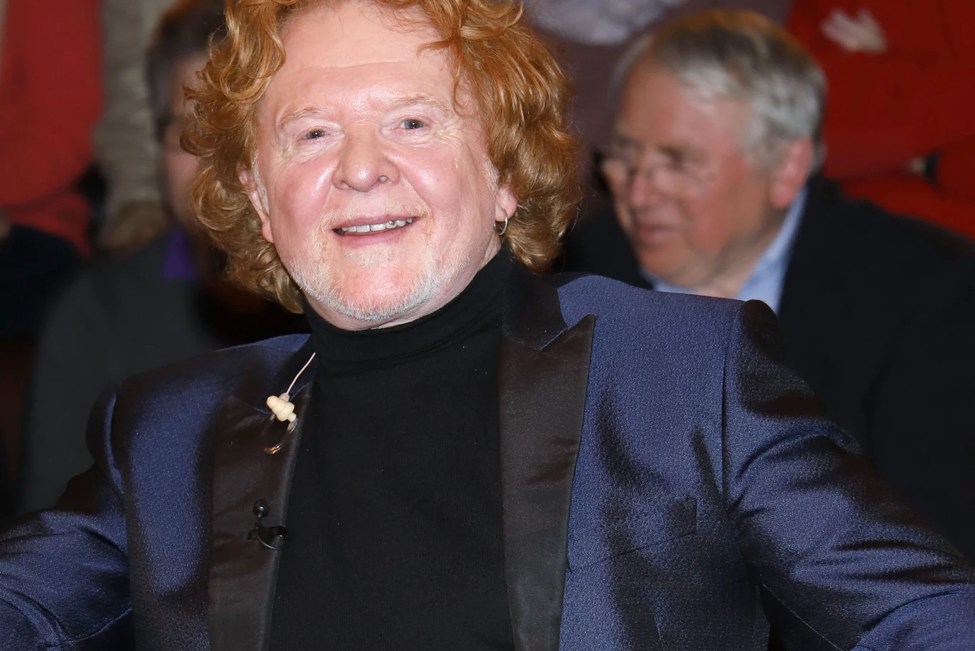 Mick Hucknall, lider Simply Red Mężczyzna z rudymi, kręconymi włosami ubrany w granatową marynarkę i czarny golf uśmiecha się patrząc w stronę aparatu. W tle widoczna rozmyta postać innego mężczyzny.