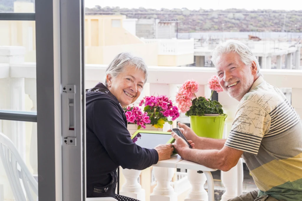 Bezpieczeństwo, dobra infrastruktura i niskie podatki dla emerytów to kluczowe czynniki decydujące o wyjeździe za granicę Starsza kobieta i mężczyzna siedzą przy stole na balkonie, uśmiechają się do kamery, wokół nich stoją doniczki z kwiatami, a na stole leżą tablet i telefon.