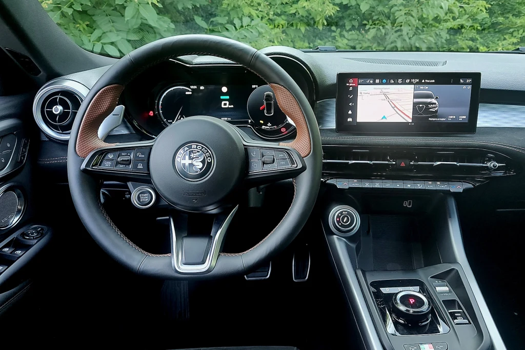 Przycisk uruchamiania silnik na kierownicy po lewej stronie, duże łopatki do zmiany przełożeń mają sugerować sportowy charakter Tonale Kierownica samochodu Alfa Romeo z widocznym logo marki, cyfrowy panel wskaźników, centralny wyświetlacz nawigacji oraz nowoczesna konsola środkowa z selektorem zmiany biegów. W tle przez szybę widoczna jest zieleń.