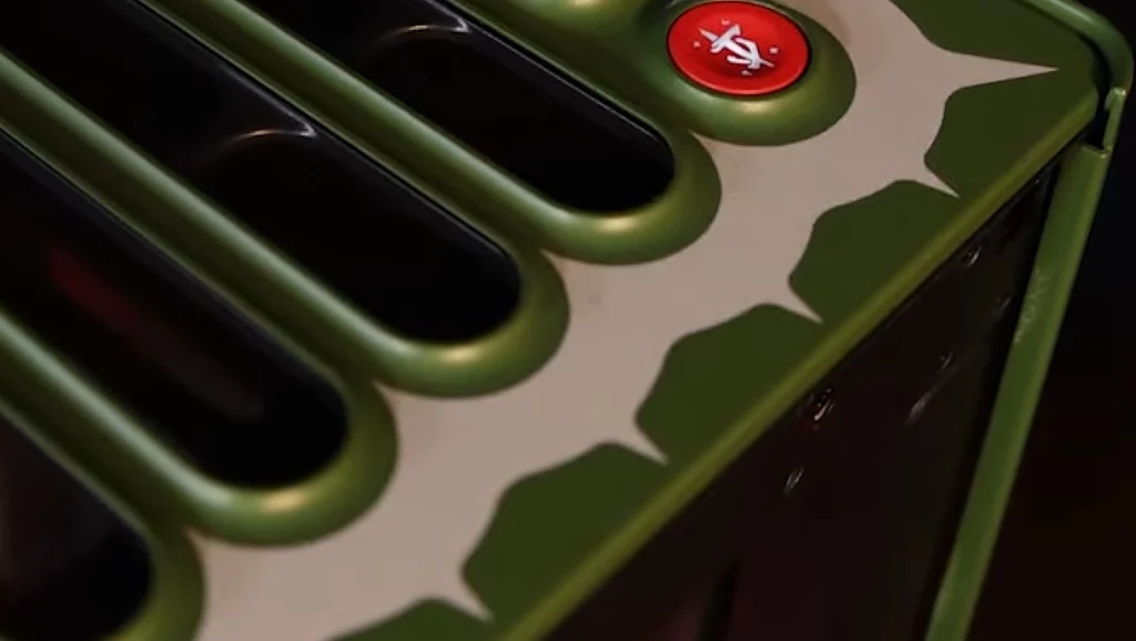 Kawałek zielonego tostera ozdobiony motywem zębów rekina oraz czerwonym przyciskiem z oznaczeniem dźwięku, przód urządzenia z wyraźnymi szczelinami na tosty.